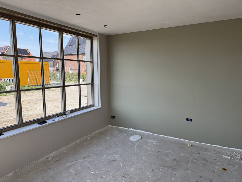 Nieuwe kamer met een groot raam met roeden, een geverfde grijsgroene wand en een betonvloer, klaar voor de volgende afwerking