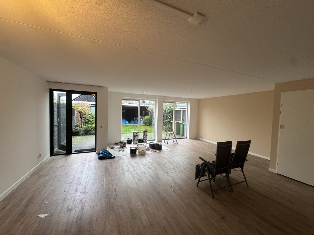 Ruime woonkamer met lichte wanden, een houten vloer en verfemmers en een trapladder bij de tuindeuren